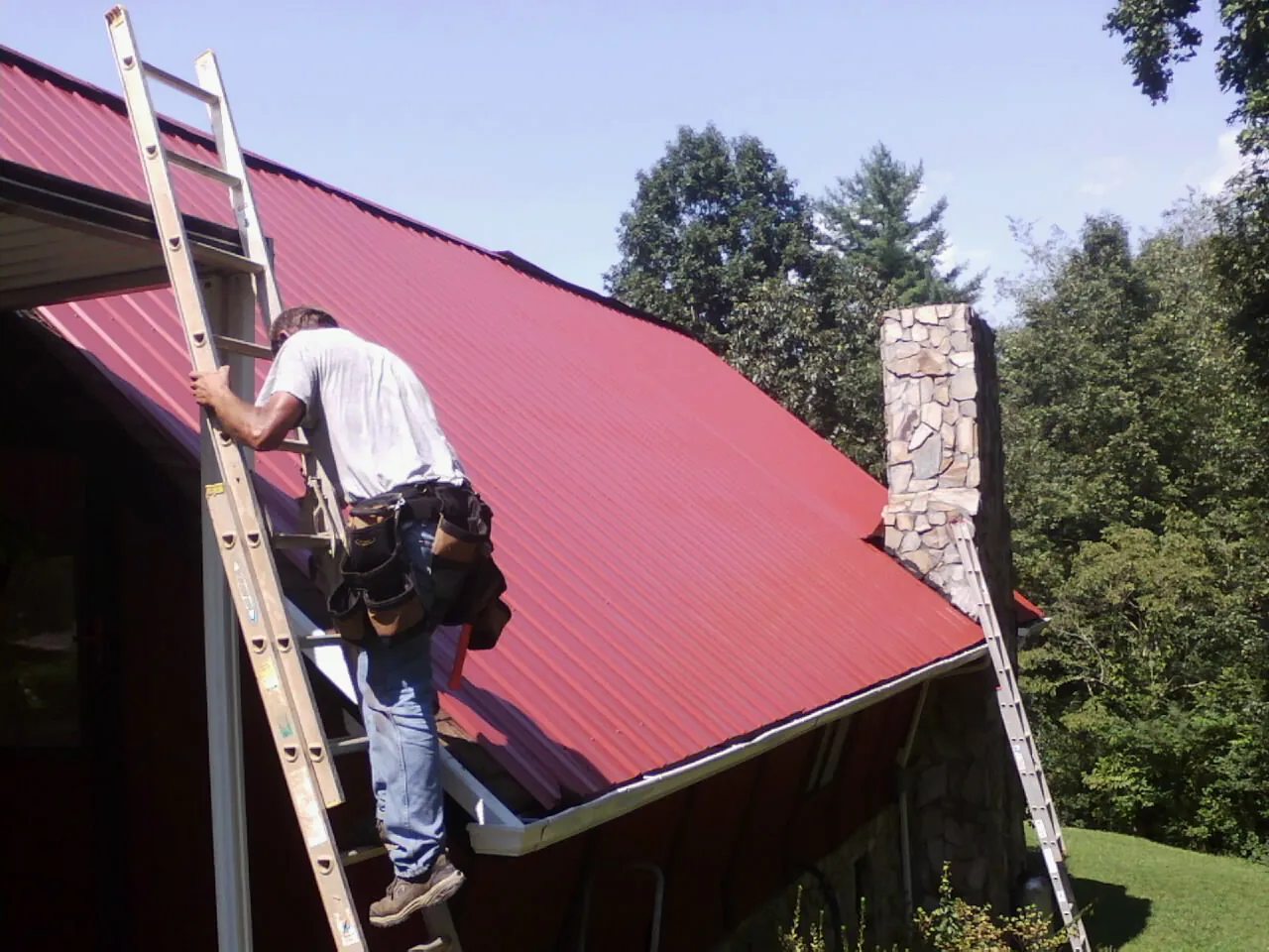 Roofer on a ladder beside a red metal roof and stone chimney — Soffit & Fascia Repair by Roofing Brothers in Mindoro